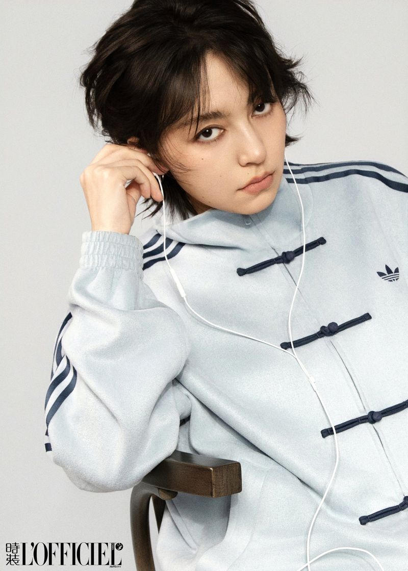 Portrait of a young Asian woman with short dark hair and light skin wearing an Adidas zip-up jacket and wired earbuds in a photo studio.