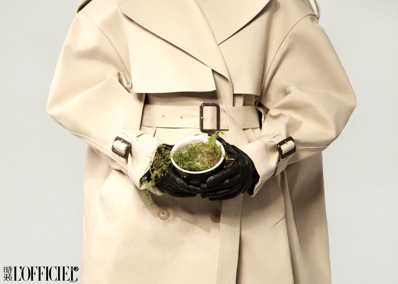 Fashion shot featuring a person in a beige trench coat holding a pot of moss against a clean background with a magazine logo.