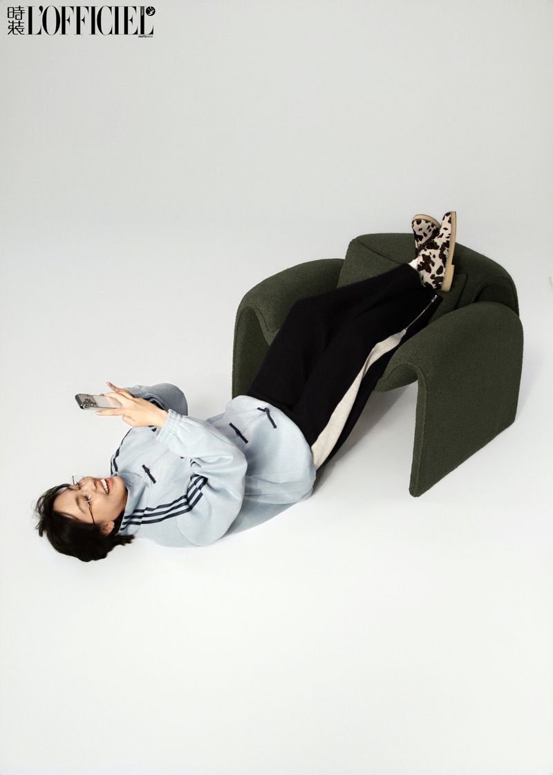 A smiling woman in athleisure wear reclines in a green chair, using a smartphone against a white backdrop in a fashion editorial.
