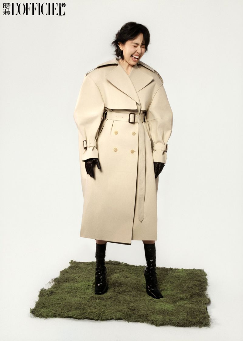 A stylish adult female wearing a beige trench coat, black gloves, and boots, laughing in a studio setting with a moss platform.