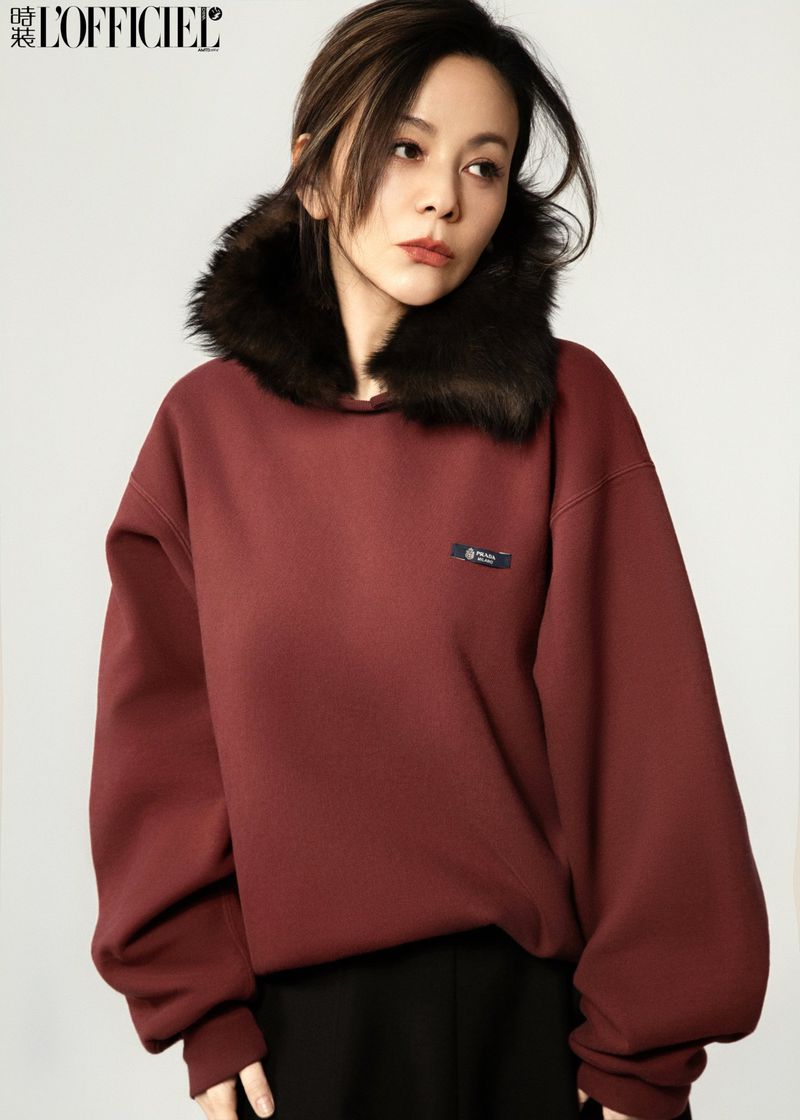 Portrait of a woman wearing a maroon sweatshirt with fur collar and subtle makeup against a neutral backdrop in a fashion editorial shot.