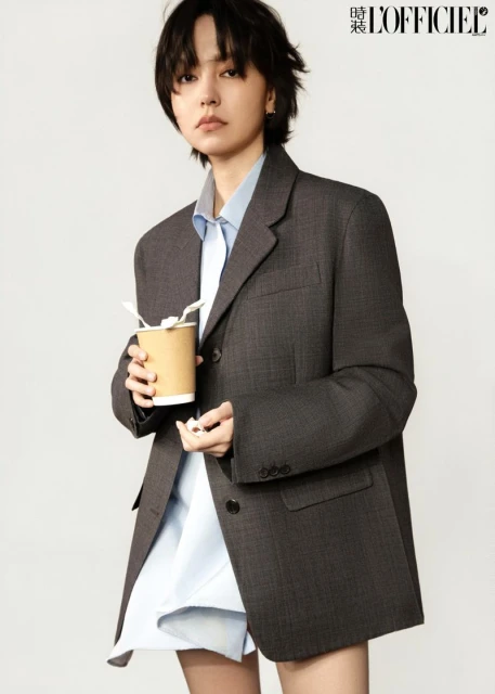 A stylish young woman in an oversized blazer holds a cup of coffee in a fashion-editorial image against a bright neutral backdrop.