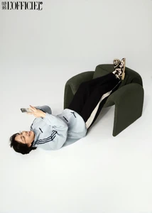 Woman in light blue track jacket and black sweatpants lying on floor looking at phone with green armchair