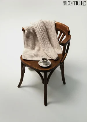 A cream textured knit blanket draped over a dark wooden chair beside a small espresso cup against a seamless white studio background.