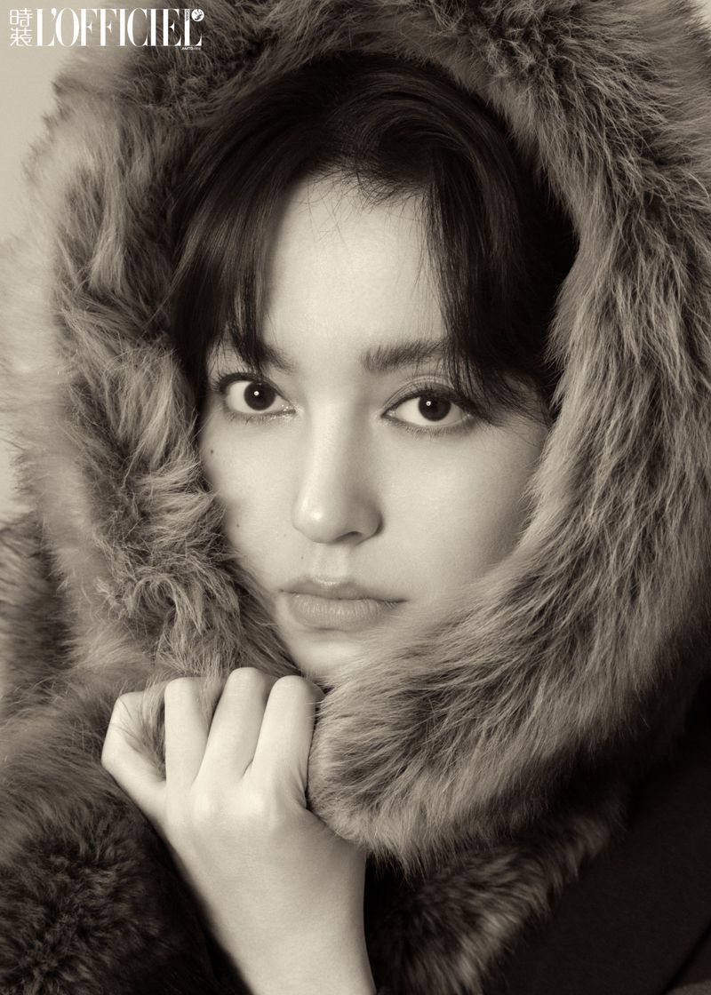 Close-up portrait of an East Asian woman wearing a plush fur hood holding the collar against a neutral background.