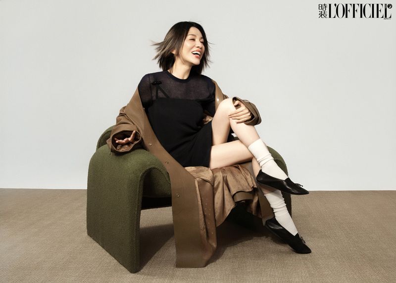 East Asian woman smiling while seated in an olive green armchair wearing a black mini dress, brown coat, and white socks.