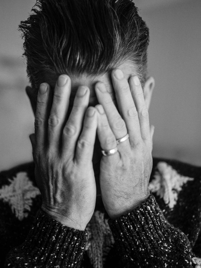 Black and white portrait of Ethan Hawke covering his face with both hands while wearing a patterned knit sweater.