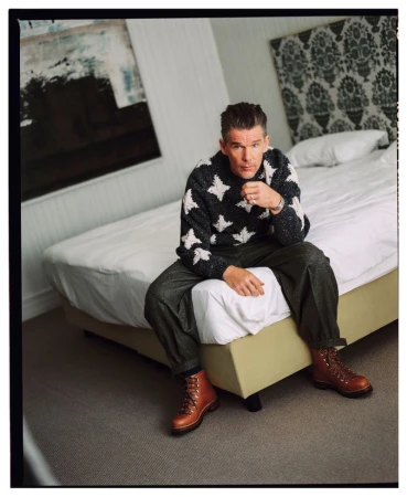 Ethan Hawke sits on the edge of a bed wearing a patterned knit sweater, dark trousers, and brown lace-up leather boots.