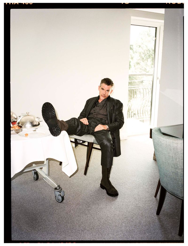 Man in dark clothing and boots poses with a foot up on a table with food service in a brightly lit room with carpet flooring.