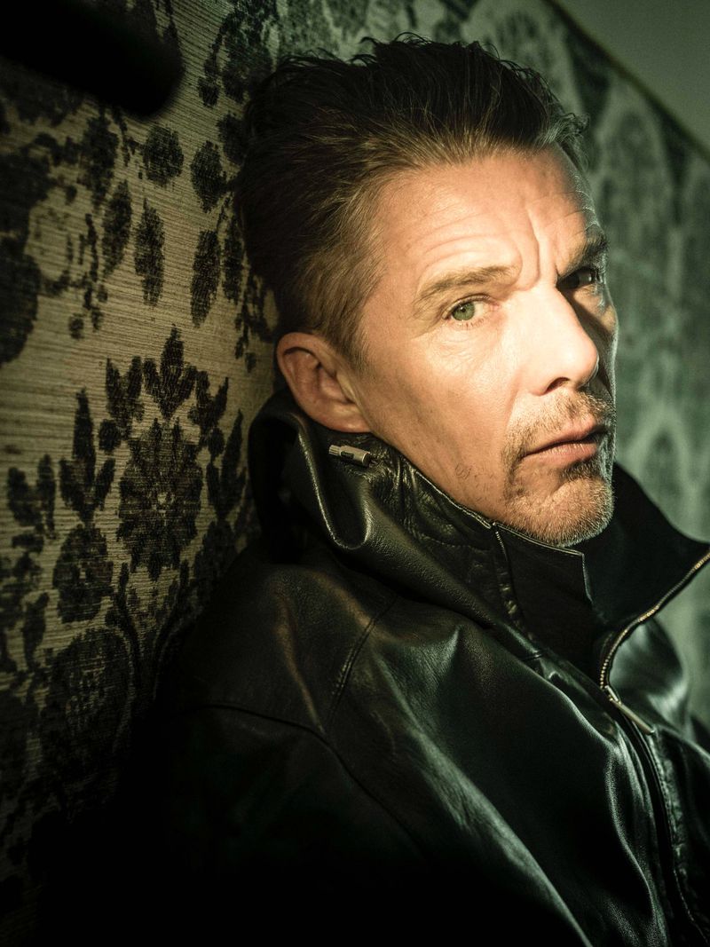Portrait of a mature adult male with a focused expression, wearing a black leather jacket, leaning against a floral patterned wall.