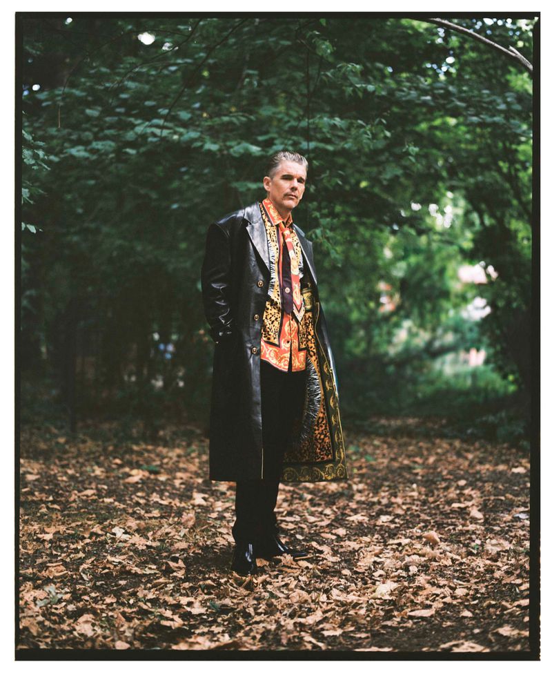 An adult male stands in a wooded park setting wearing a long black leather coat over a patterned orange shirt, styled in a fashion editorial.