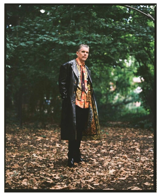 An adult male stands in a wooded park setting wearing a long black leather coat over a patterned orange shirt, styled in a fashion editorial.