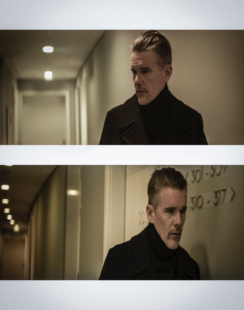 Close-up of a man with dark hair and a black turtleneck and coat in an interior hallway under dim lighting, looking away.