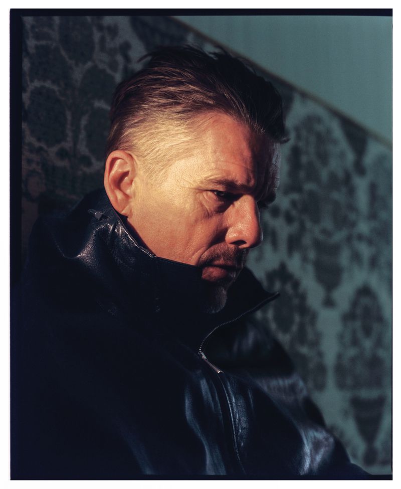 Portrait of adult male with short hair and goatee wearing a dark jacket against a blue patterned wallpaper backdrop in a photo studio.