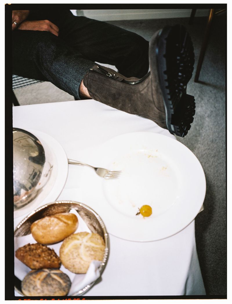 Person in grey trousers and brown suede boot rests foot on a table, near a plate with bread rolls and a single grape.