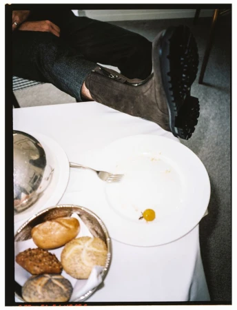 Person in grey trousers and brown suede boot rests foot on a table, near a plate with bread rolls and a single grape.