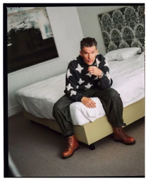 Ethan Hawke sits on the edge of a bed wearing a patterned knit sweater, dark trousers, and brown lace-up leather boots.