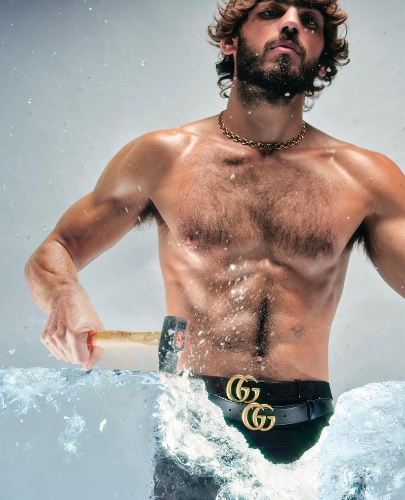 Muscular man poses shirtless behind ice block, breaking it with hammer. Gold chain necklace and Gucci belt visible against light background.