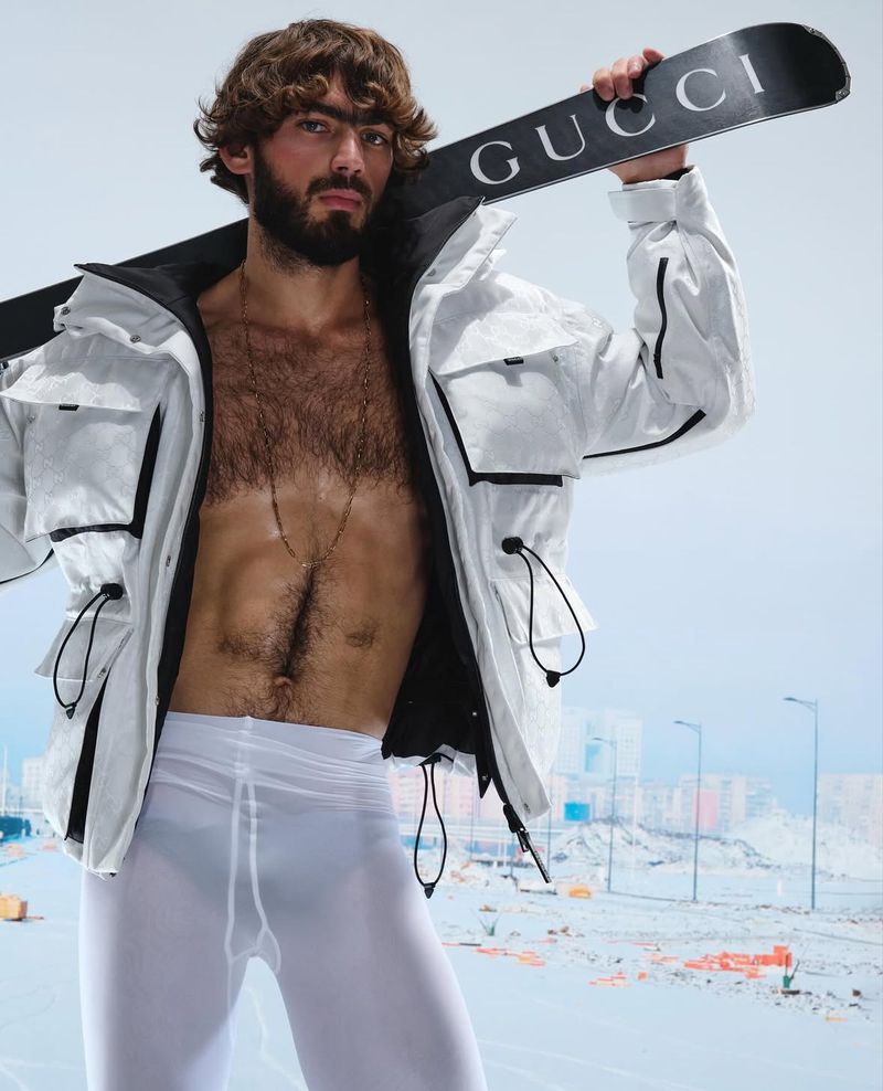 A man with a beard poses with a Gucci snowboard, wearing a white jacket and white tights, against a blurred background of buildings.