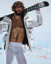 Man with beard holding a Gucci branded ski on his shoulder wearing an open white jacket and white tights against an outdoor snow background