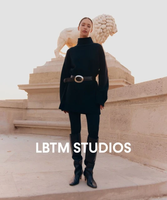 A woman models black tunic, leggings, and heeled boots in an outdoor fashion shoot with a stone lion statue in the background.