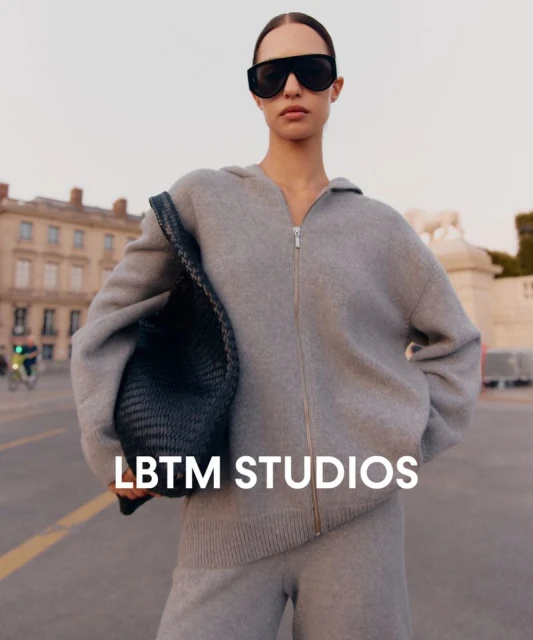Woman in grey hooded sweatshirt and sweatpants stands on a street with European buildings, wearing sunglasses and holding a black tote bag.