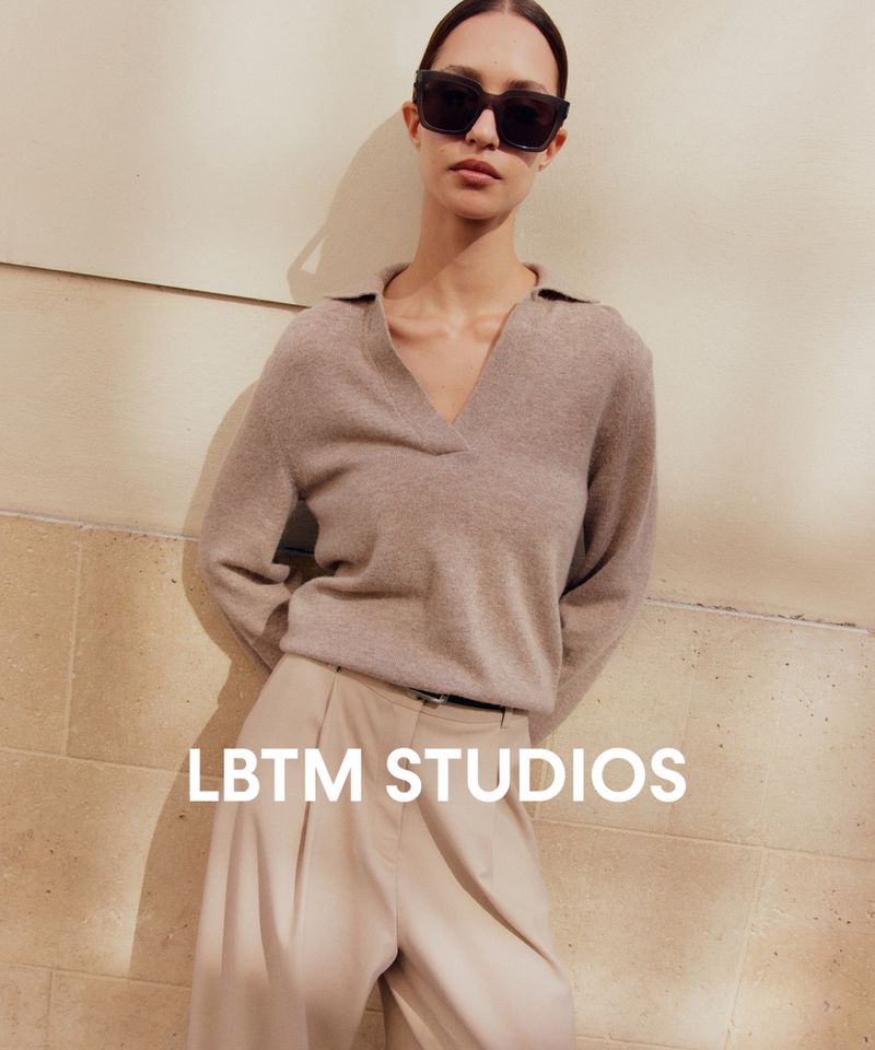 Woman wearing black sunglasses, a taupe knit sweater, and pleated beige trousers leaning against a stone wall.