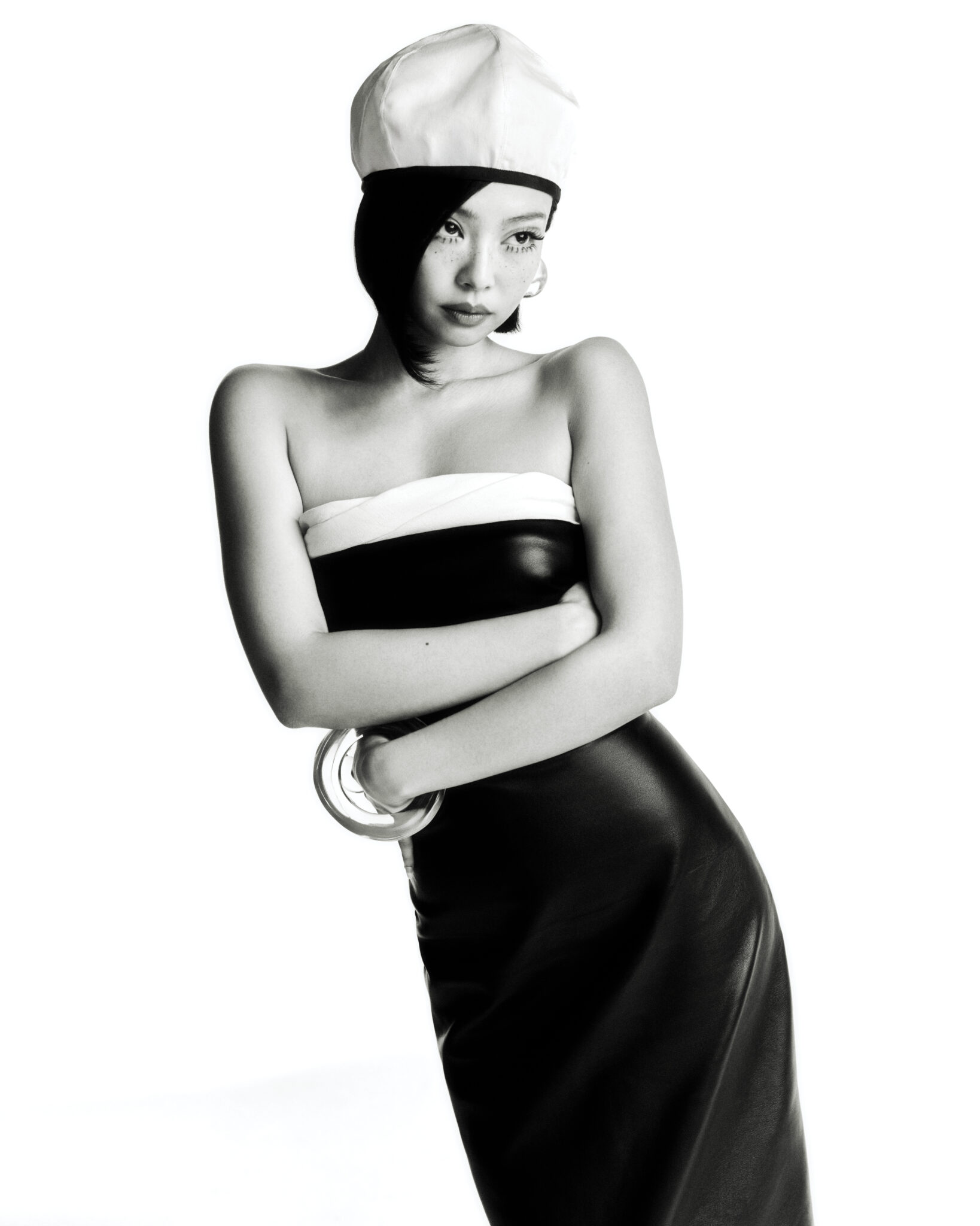 Striking black-and-white studio shot of a stylish adult female model wearing a black dress and white hat, set against a bright backdrop.