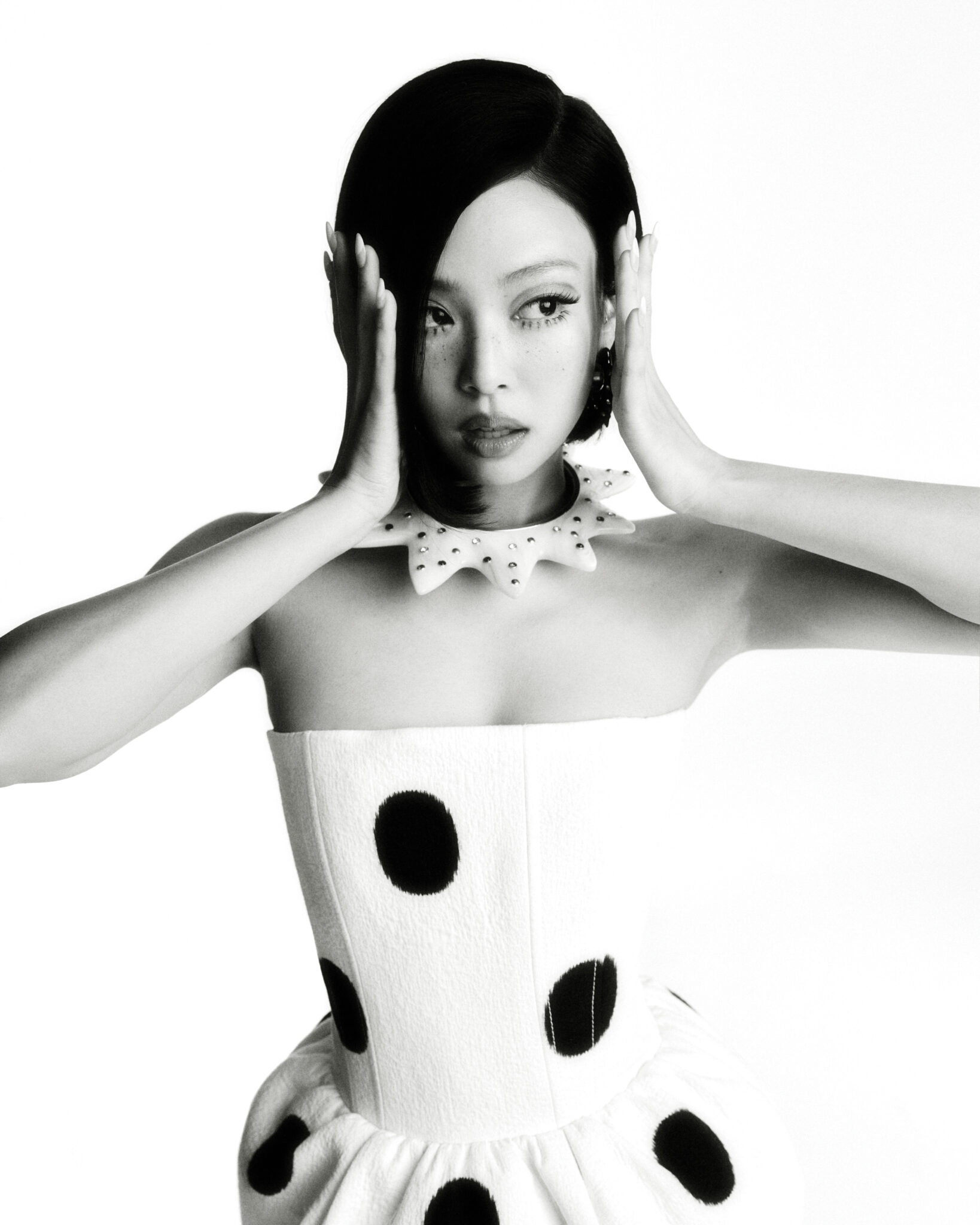 Black-and-white portrait of a stylish woman with dark hair in a polka dot dress against a white backdrop, hands near face.