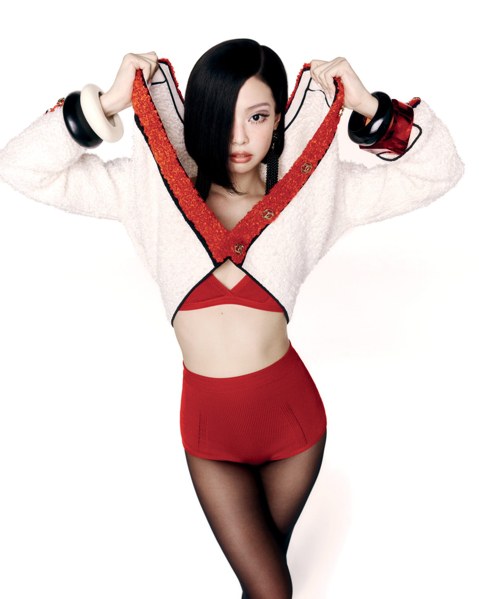 A fashionable young adult woman with short black hair poses in a stylish white jacket with red trim, red top, red shorts, and black tights.