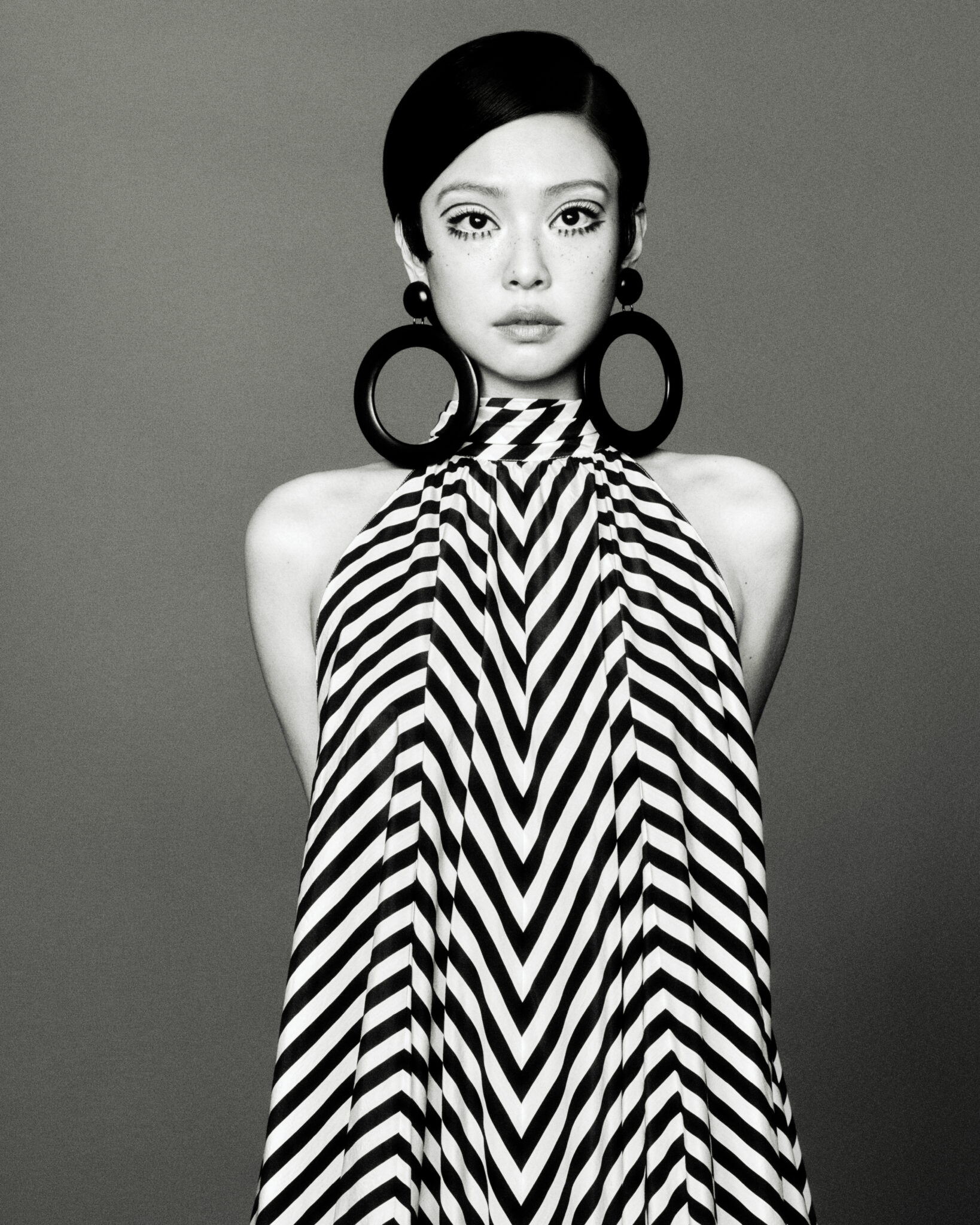 Black and white portrait of an Asian woman with dark hair, large hoop earrings, and graphic eyeliner, wearing a black and white striped dress.