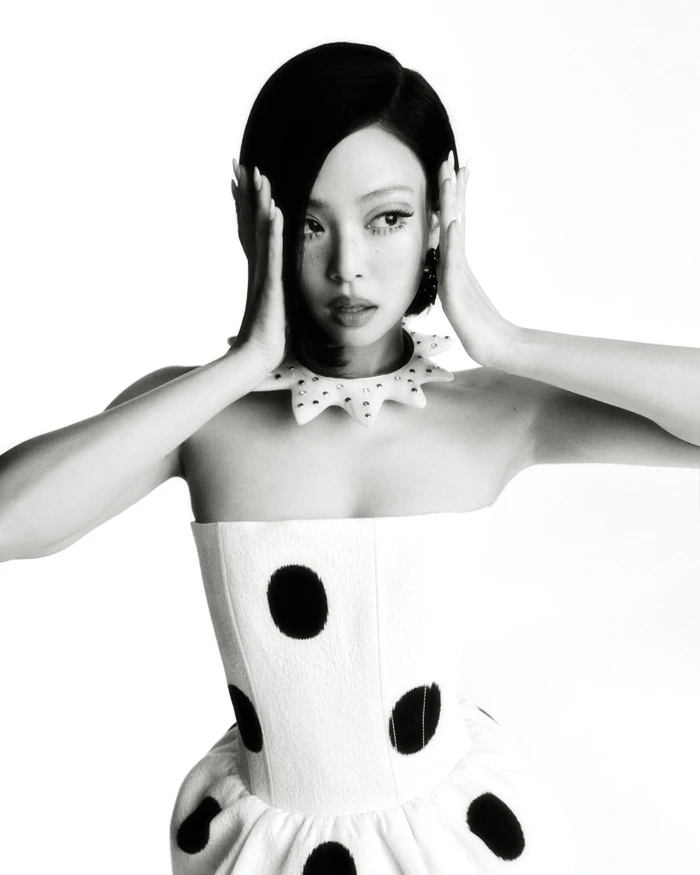 Jennie wearing a white polka dot strapless dress and spiked collar with hands framing her face in a high-contrast portrait for V Magazine