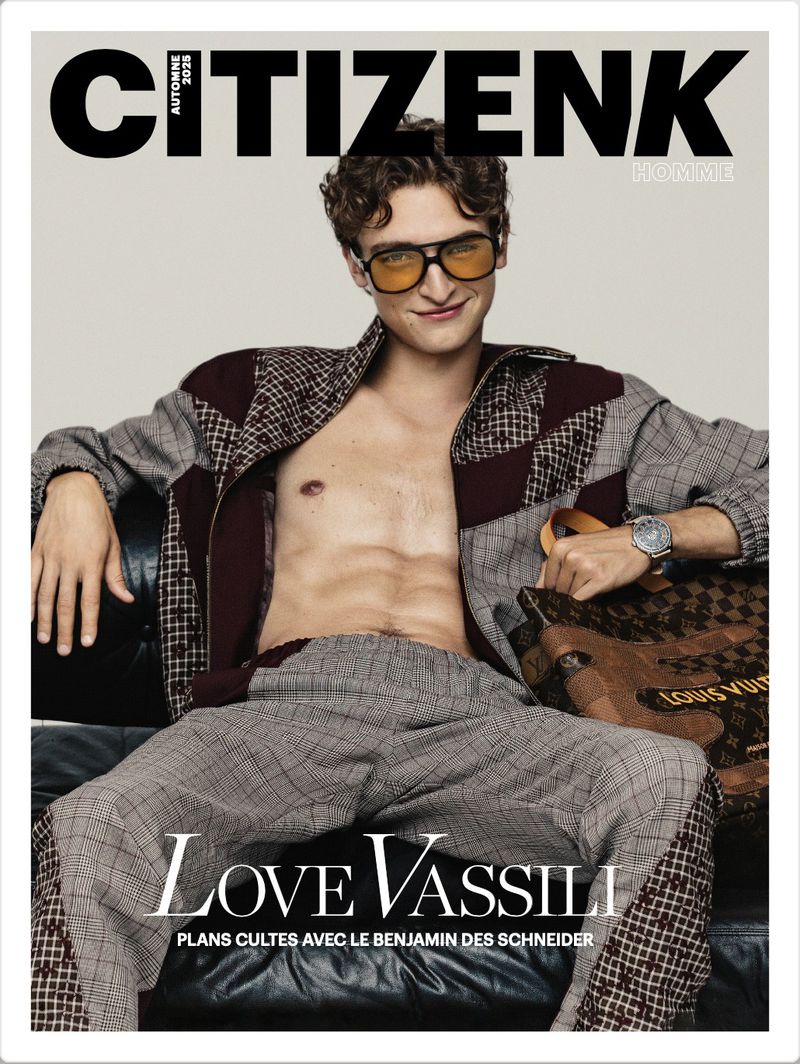 Actor Vassili Schneider wearing amber-tinted sunglasses and an open-front patterned jacket seated on a dark leather sofa