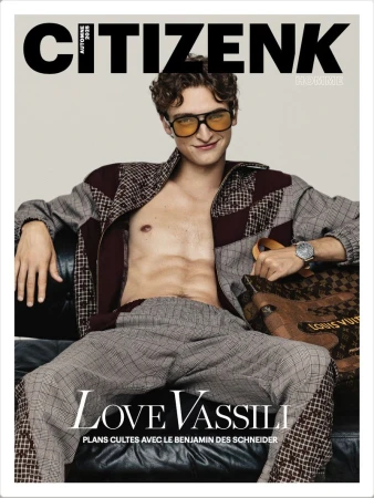Actor Vassili Schneider wearing amber-tinted sunglasses and an open-front patterned jacket seated on a dark leather sofa