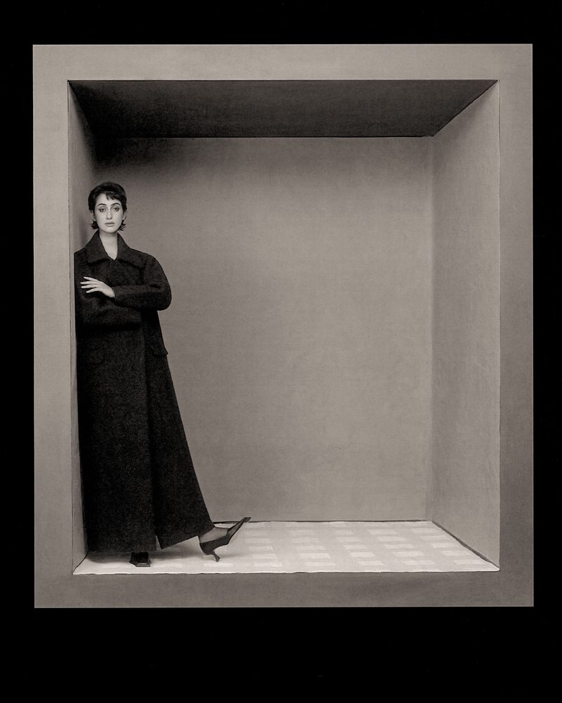 Monochrome fashion portrait of a slender woman in a long coat, leaning against the corner inside a gray cubical structure. 