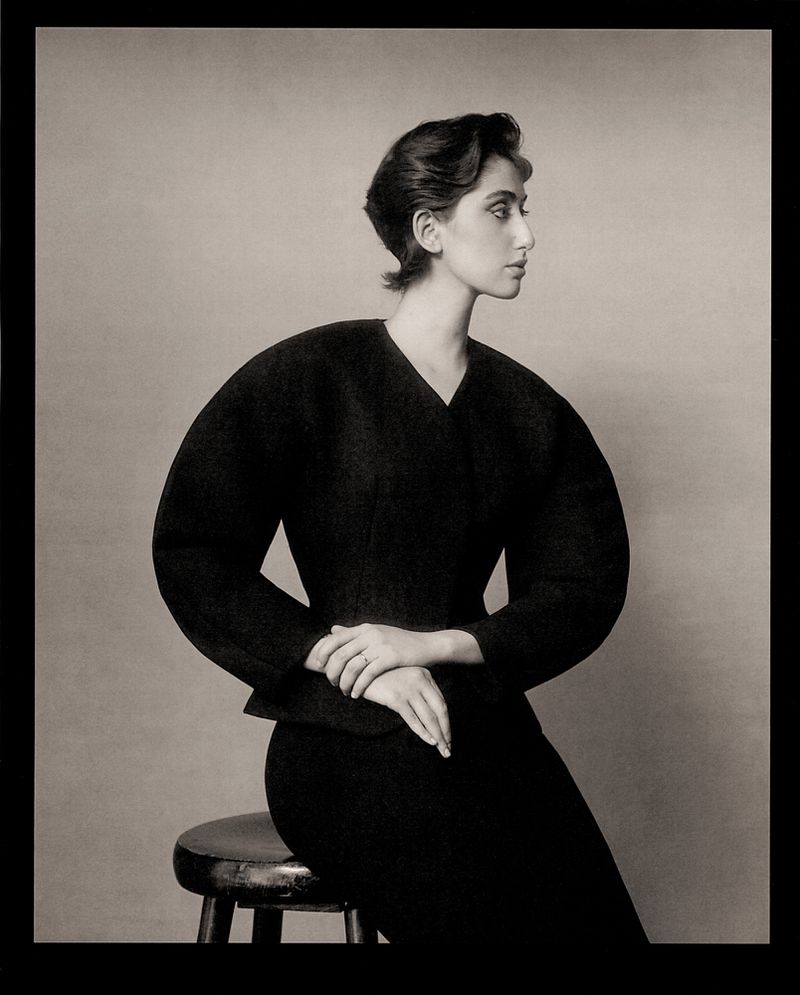 Profile shot of a woman in a black suit jacket and skirt sitting on a stool in front of a plain grey background in a studio setting.
