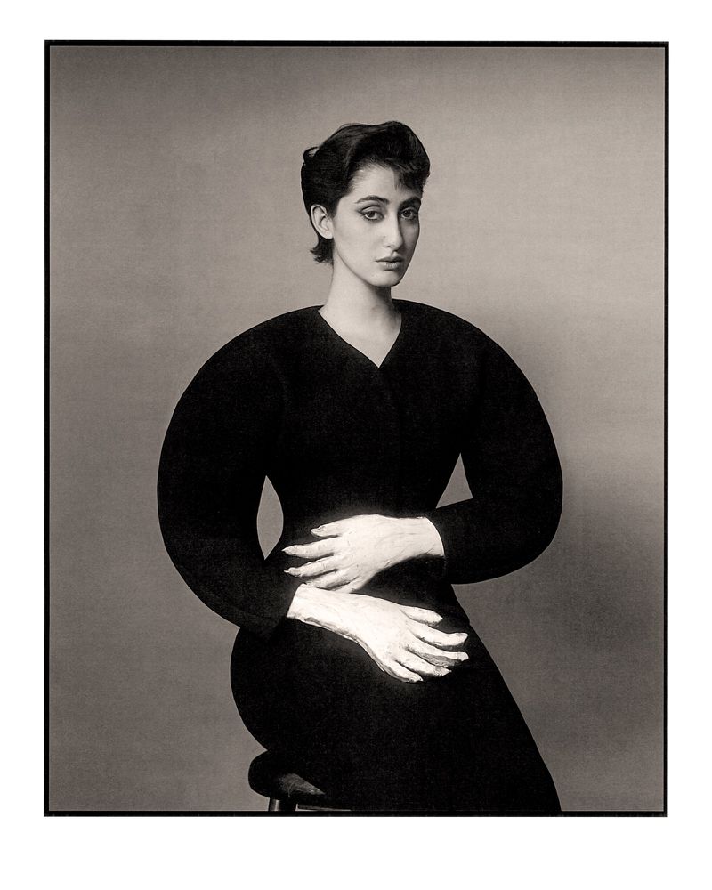 Elegant woman in a black dress and white gloves sits in a studio, posing gracefully against a neutral backdrop, showcasing classic sophistication.