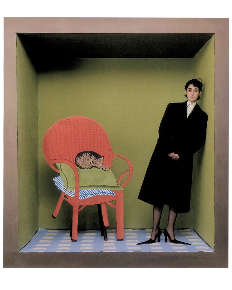 A woman in a black coat poses in a grey box with a red chair and a kitten, with a studio photography, creating an artistic and staged scene.