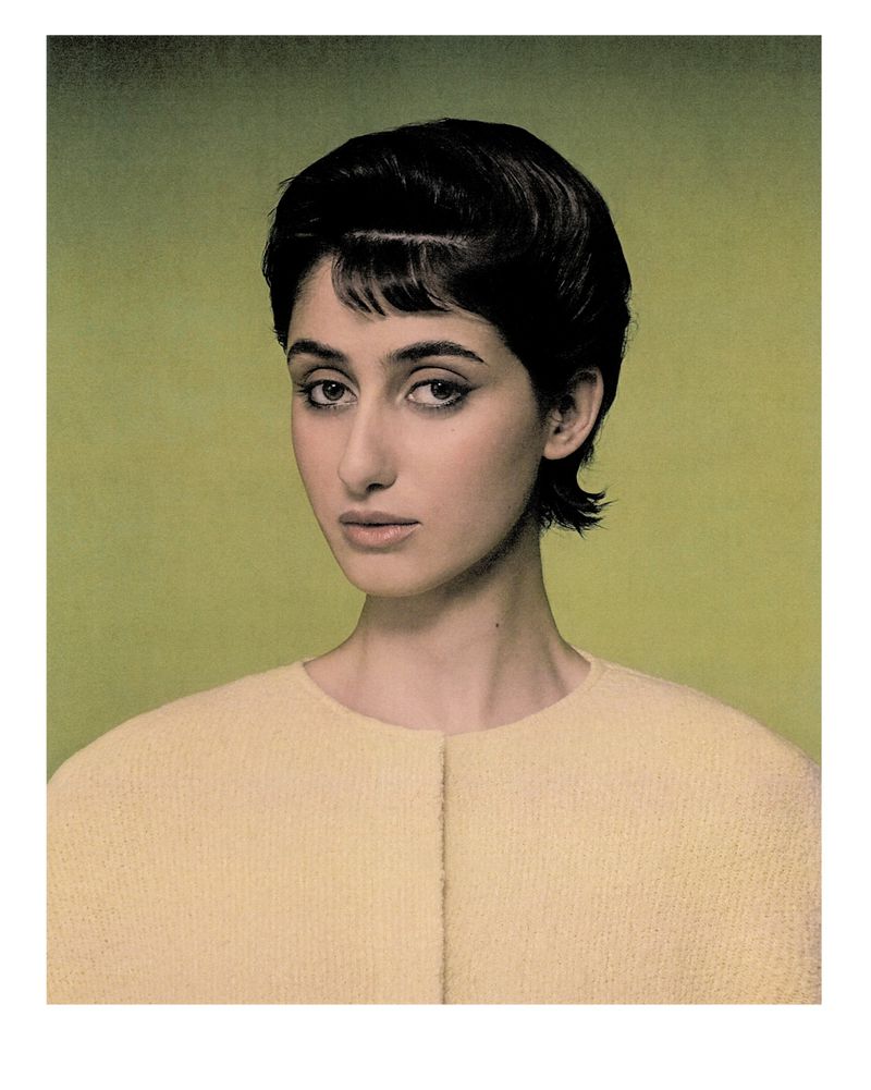 Portrait of a woman with dark hair and light skin, wearing a cream-colored top, against a muted green background. Beauty shot, close-up.