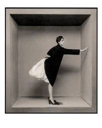 Model in a black coat and voluminous white bubble skirt posing inside a recessed rectangular box structure against a neutral backdrop.