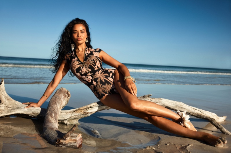 Woman in tropical print swimsuit reclines on driftwood at the beach against a blue sky and ocean backdrop on a sunny day.