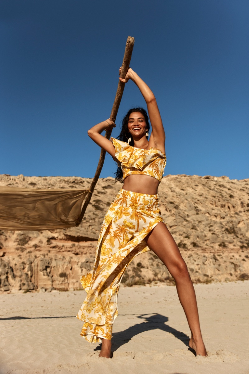 A woman in a yellow patterned crop top and skirt smiles while holding a wooden pole on a sandy beach with a desert backdrop under a blue sky.