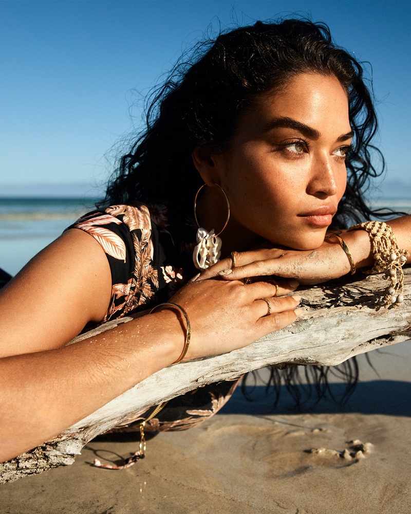 Shanina Shaik leaning on a driftwood log on a sandy beach wearing a tropical print top and hoop earrings for Seafolly.