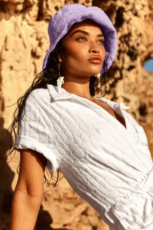 Shanina Shaik in a white textured romper and lilac bucket hat against a rocky outdoor backdrop