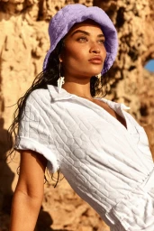 Shanina Shaik in a white textured romper and purple bucket hat against a rocky coastal background
