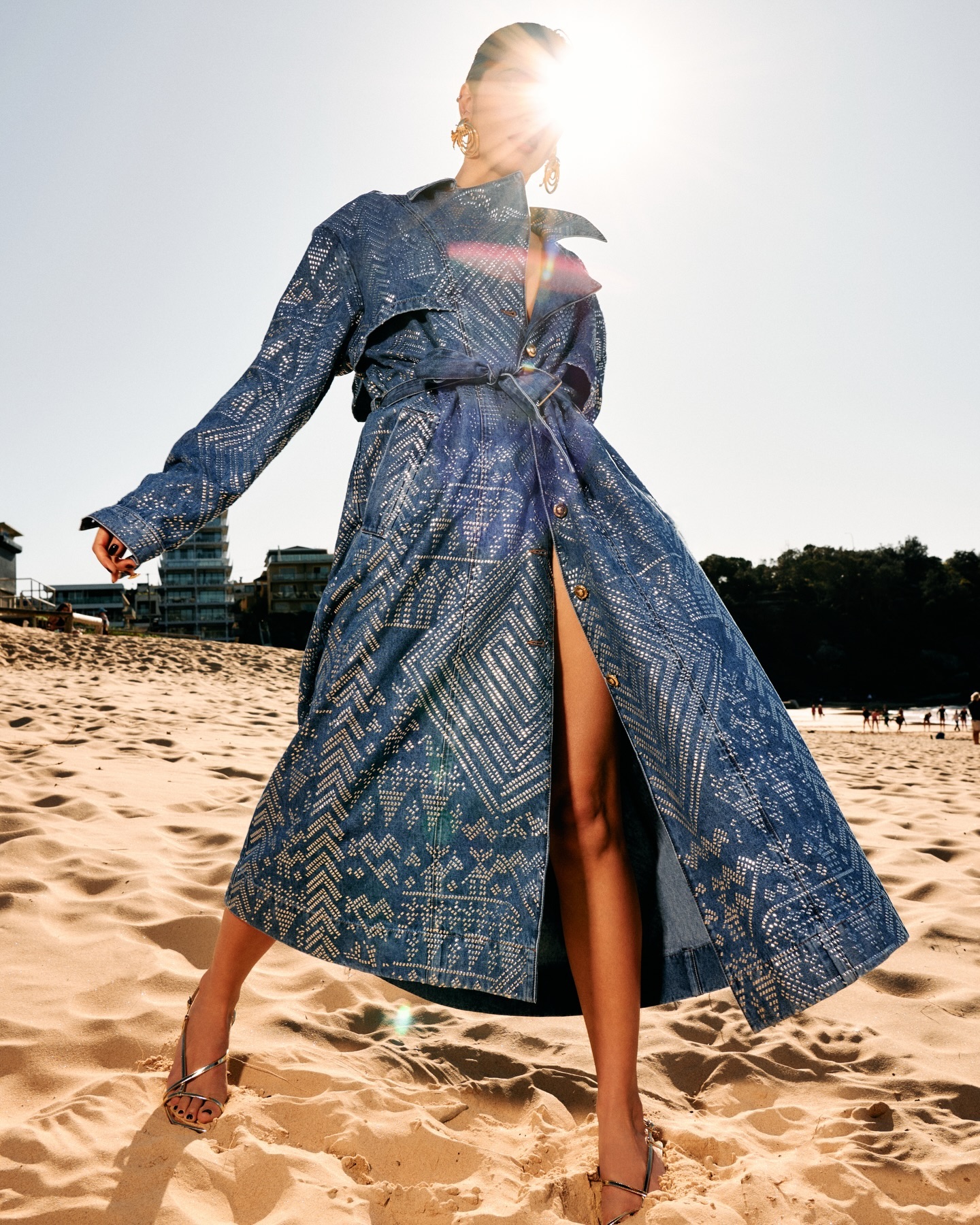 Model wearing a printed blue denim trench coat standing on a sandy beach under bright sunlight.