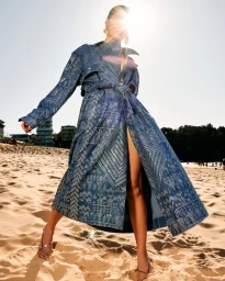 Model wearing a printed blue denim trench coat standing on a sandy beach under bright sunlight.