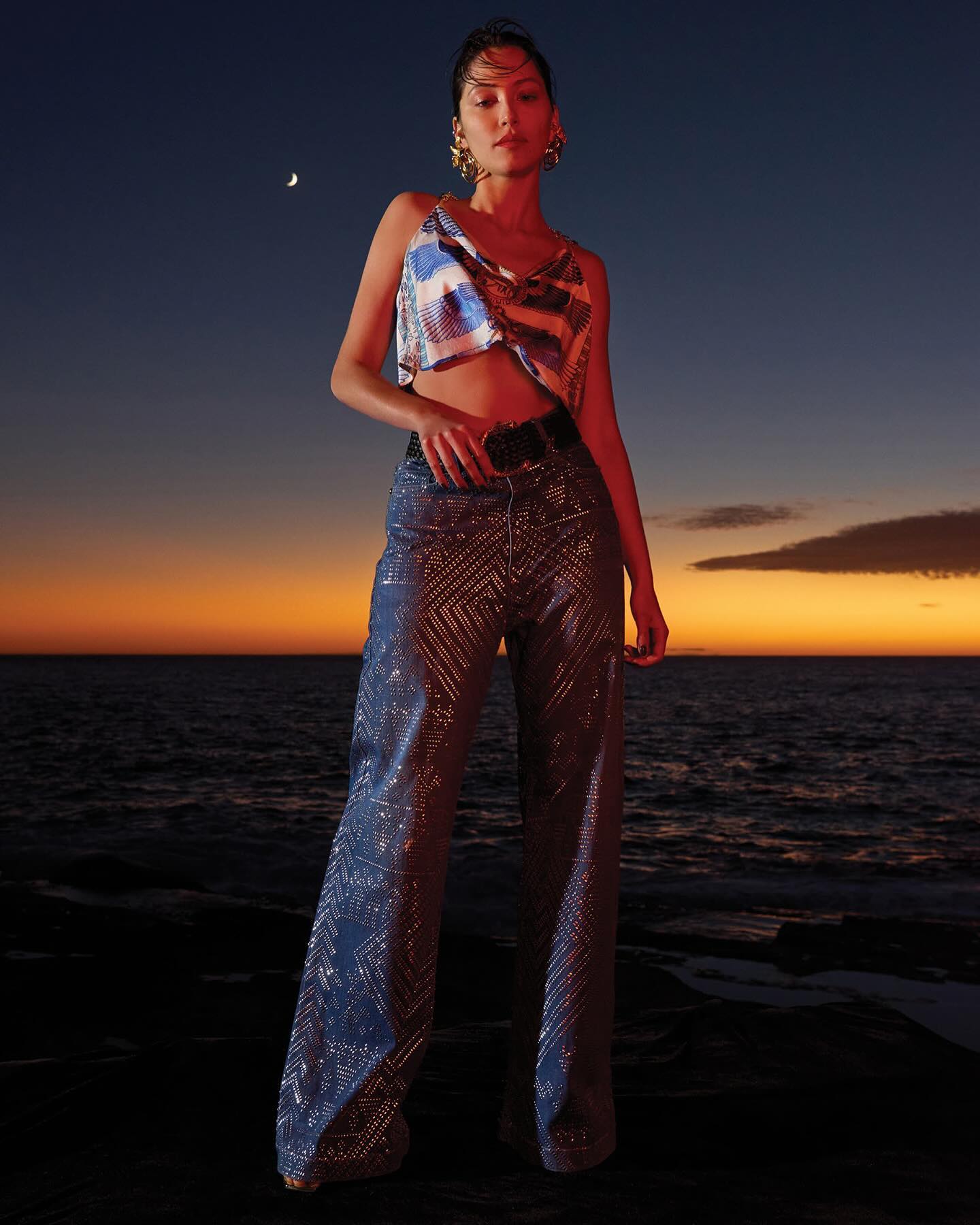 Fashionable young woman posing outdoors at dusk in a crop top and patterned pants, showcasing a blend of modern style and evening ambiance.