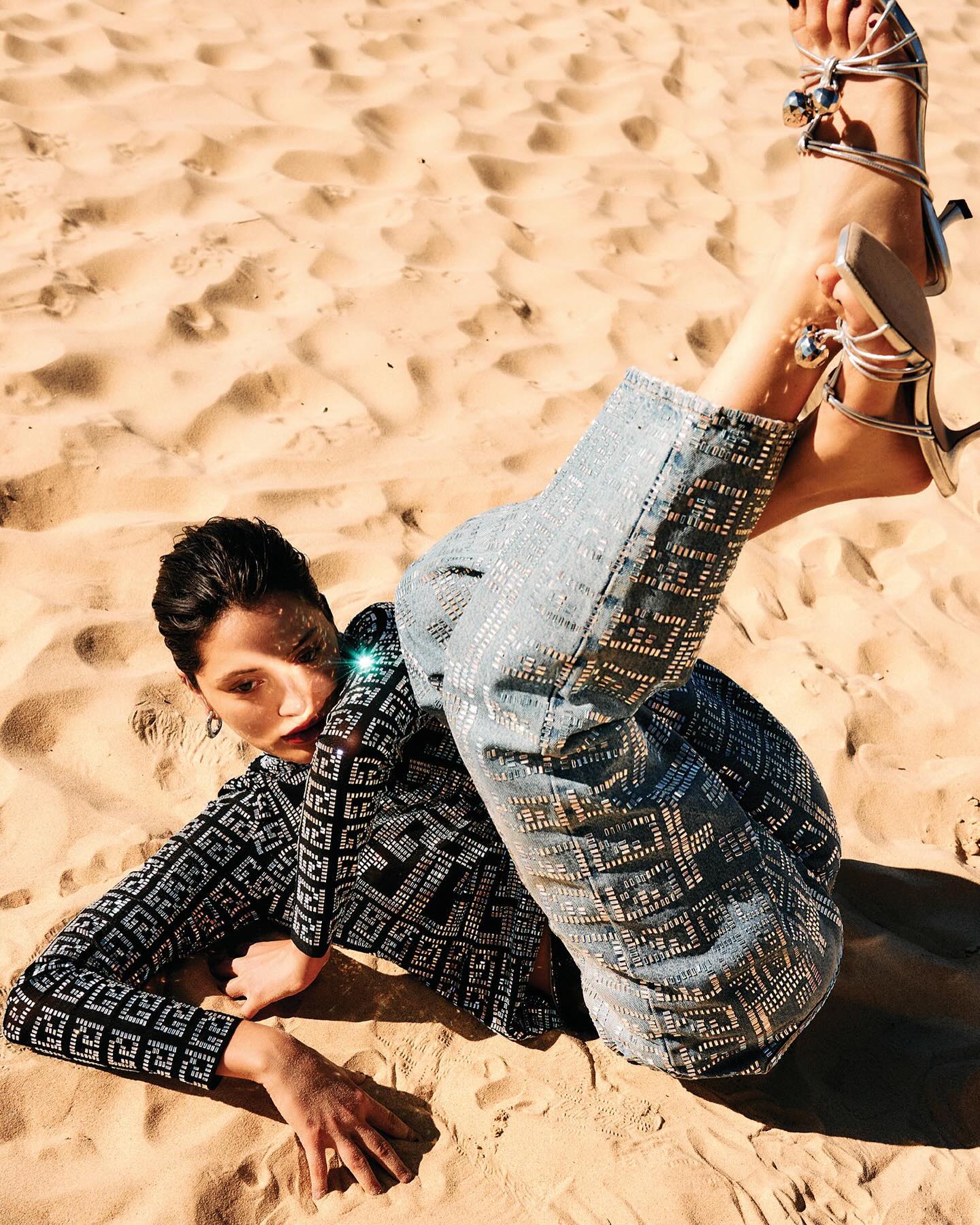 Model lying on sandy beach wearing patterned top, embellished jeans, and silver strappy heels with legs raised
