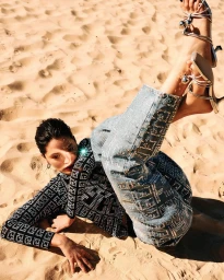 Model lying on sandy beach wearing patterned top, embellished jeans, and silver strappy heels with legs raised